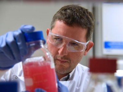 A male scientist in lab coat and goggles inspects a glass full of red liquid. 